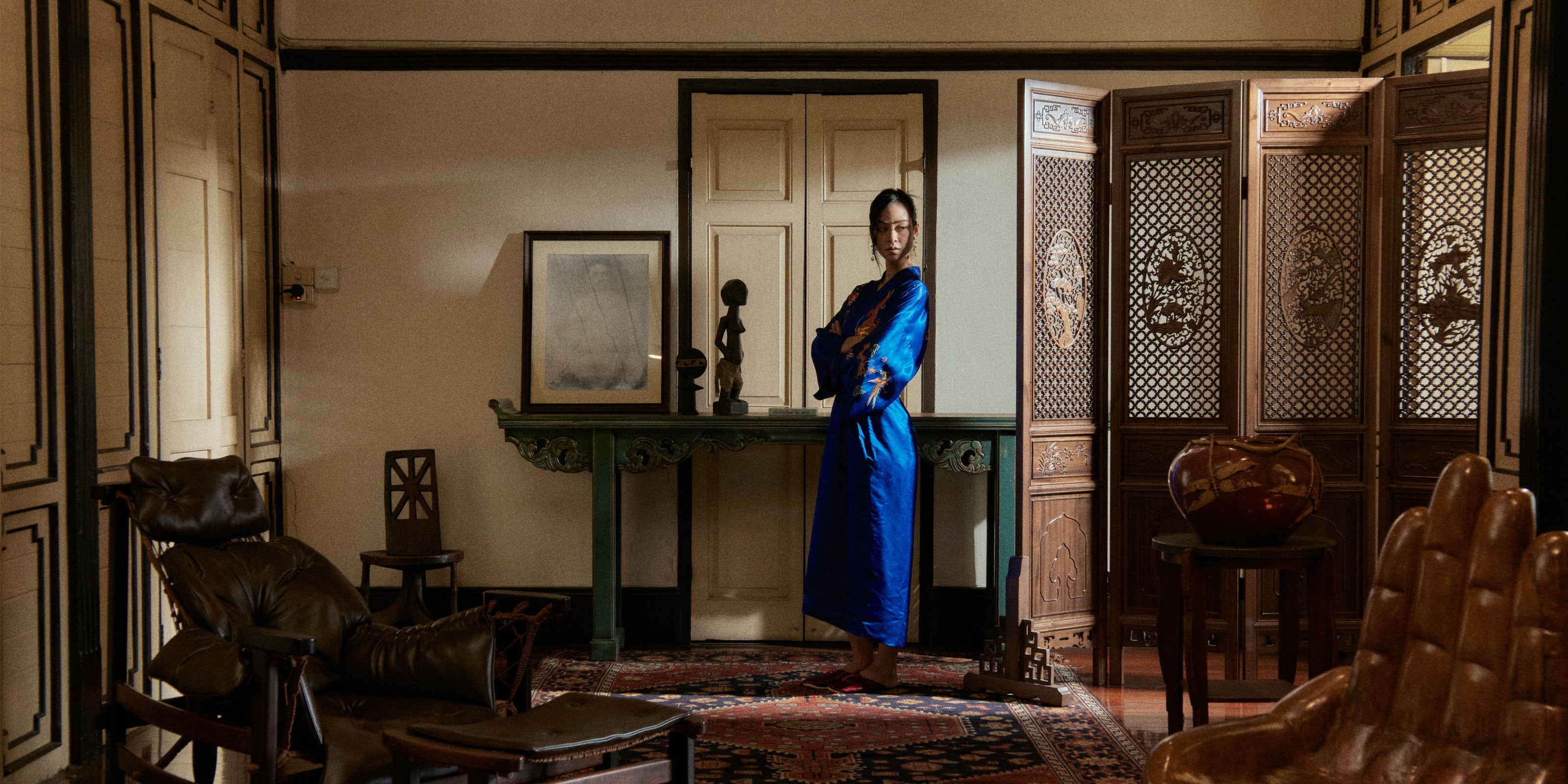 A woman in a blue robe stands confidently in a dimly lit vintage room with leather chairs, ornate wooden divider, and a Persian rug, exuding elegance and mystery.