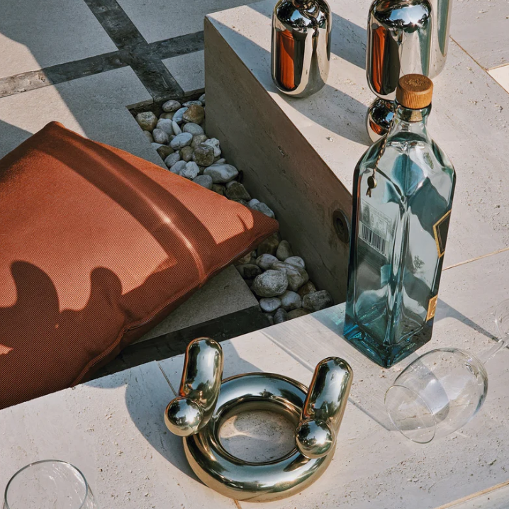 Outdoor setting with a brown cushion, silver decorative object, and tall glass bottles on a patio. Light creates soft shadows, conveying a relaxed vibe.